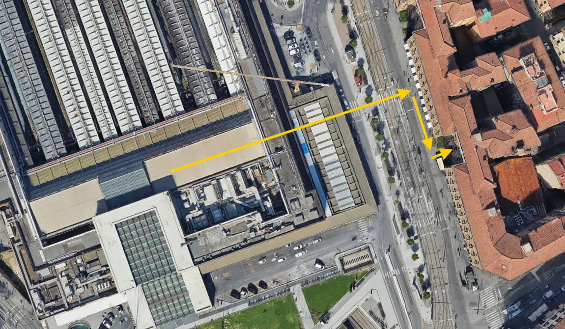 Vista aerea di una stazione ferroviaria con binari e tetti visibili sulla sinistra e un quartiere residenziale con edifici dai tetti rossi sulla destra. Sono presenti delle frecce gialle che indicano un percorso dalla stazione verso un punto specifico del quartiere.